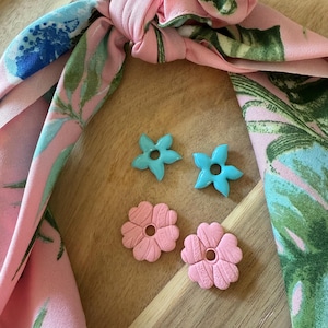 Puede incluir: Una cinta para el pelo con estampado floral rosa con dos pendientes en forma de flor turquesa y dos pendientes en forma de flor rosa. La cinta para el pelo tiene un estampado de hojas tropicales en rosa, verde y azul.