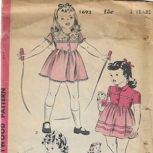 May include: A vintage Hollywood sewing pattern envelope for a child's dress, labeled 1693 and 3 Years. The illustration shows four variations of a dress with short sleeves, button fronts, and collars, worn by children playing with toys.