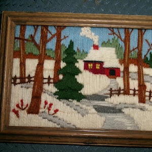 May include: A framed yarn embroidery depicting a winter landscape with a red cabin featuring a white snow-covered roof and chimney smoke. The scene includes a green pine tree, bare brown trees, a wooden fence, and a path set against a blue sky.