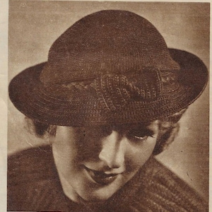 May include: A sepia-toned image of a brown hat with a rounded crown and a wide brim. The hat features a decorative bow detail on the front. The hat is made of woven material.