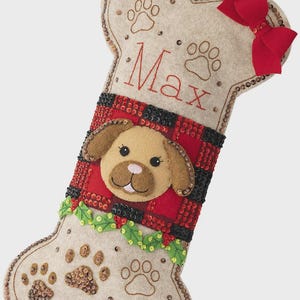 May include: A bone-shaped Christmas stocking made of beige felt with a red and black buffalo plaid center panel. It features an embroidered dog face, a red felt bow, green holly leaves, and sequin-accented paw prints. The name Max is embroidered in red.