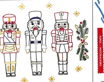 May include: An embroidery transfer pattern featuring three nutcracker figures in red, gold, and black outlines, surrounded by green pine sprigs with red berries and yellow starbursts. The text reads Aunt Martha's and #SPEC - NUTCRACKERS.