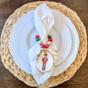 May include: A white napkin is held by a beaded napkin ring with red, green, and natural wood beads. A painted oyster shell with a gold rim features a nutcracker design, resting on white plates and a woven placemat.
