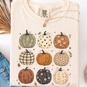 May include: An ivory crew neck t-shirt featuring a graphic print of nine pumpkins in various patterns and colors, including floral, plaid, heart, and dotted designs. The shirt has a Comfort Colors brand label at the inner neckline.