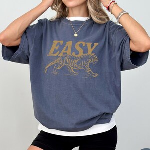 May include: A denim blue t-shirt with the word "EASY" in gold lettering above a gold tiger graphic. The shirt has short sleeves and a white undershirt is visible. The model is wearing black shorts.