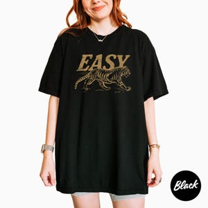 May include: Black t-shirt with the word "EASY" in a gold, vintage font above a gold tiger illustration. The tiger is walking to the right. The t-shirt is oversized and has short sleeves.