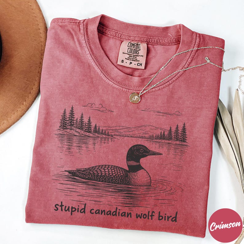 May include: Crimson-colored t-shirt with a graphic of a loon on a lake, surrounded by trees and mountains. The text "stupid canadian wolf bird" is printed below the image. A silver necklace with a pendant is draped on the shirt.