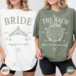 May include: Two oversized t-shirts, one white and one moss green. The white shirt says "BRIDE" with a tiara graphic and "Happily Ever After." The green shirt says "THE BACH Club" with a disco ball graphic and "Kylie's Bachelorette Party."