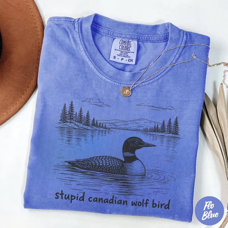 May include: A blue Comfort Colors t-shirt with a black graphic of a loon swimming in water, with a mountain and tree line in the background. The text "stupid canadian wolf bird" is printed below the graphic. A gold necklace is draped on the shirt.