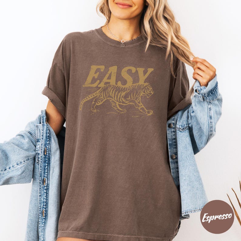 May include: Brown t-shirt with the word "EASY" in gold lettering above a gold tiger graphic. The shirt has short sleeves and a relaxed fit. The model is wearing a denim jacket.