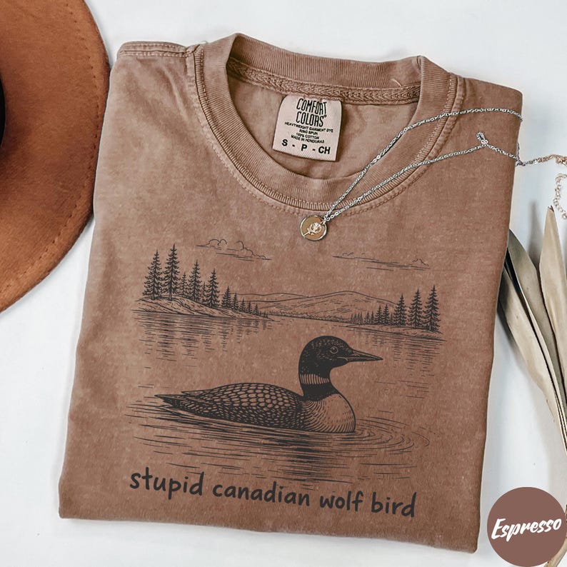 May include: Espresso-colored t-shirt with a graphic of a loon on a lake, with trees and mountains in the background. The text "stupid canadian wolf bird" is printed below the graphic. A silver necklace is draped across the shirt.