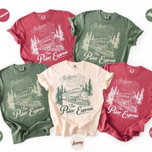 May include: Five short-sleeve crew neck t-shirts in crimson, moss, and ivory colors. Each shirt features a white or dark green graphic print of a steam locomotive train, pine trees, clouds, and the text Believe and The Polar Express.