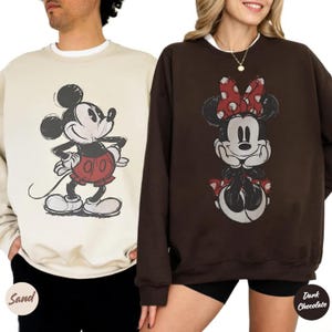 May include: Two crew neck sweatshirts with Disney character graphics. One is sand-coloured with a Mickey Mouse design in red shorts. The other is dark chocolate with a Minnie Mouse design wearing a red bow.