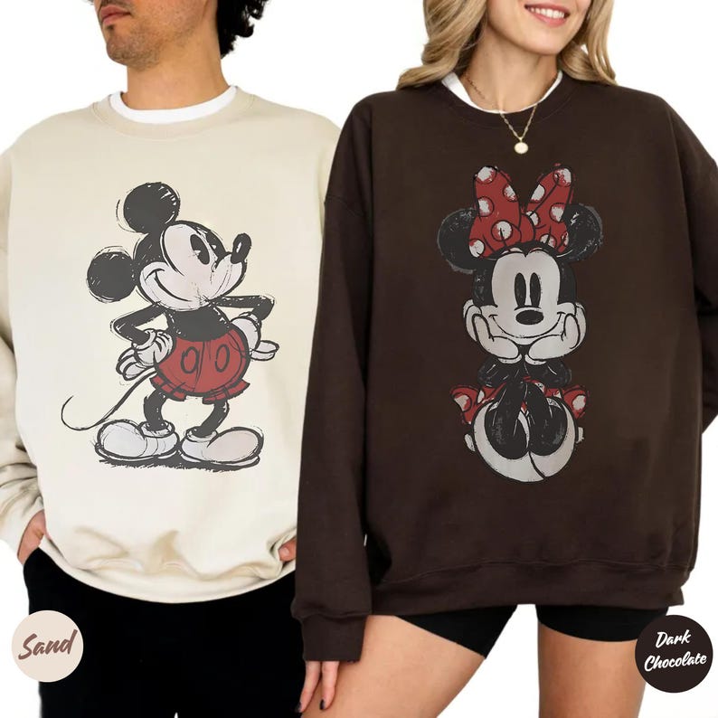 May include: Two crew neck sweatshirts with Disney character graphics. One is sand-coloured with a Mickey Mouse design in red shorts. The other is dark chocolate with a Minnie Mouse design wearing a red bow.