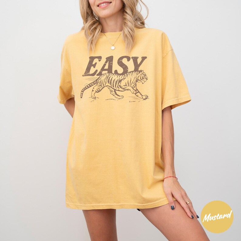 May include: Mustard yellow t-shirt with the word "EASY" in brown block letters above a brown tiger graphic. The shirt is a relaxed fit and has short sleeves.