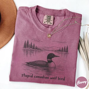 May include: Berry-colored t-shirt with a graphic of a loon on a lake, with a forest and mountain backdrop. The text "stupid canadian wolf bird" is printed below the image. A gold necklace is draped across the shirt.