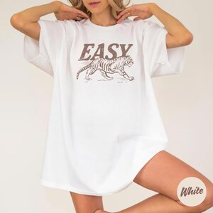May include: White t-shirt with the word "EASY" in brown block letters above a brown tiger graphic. The tiger is walking to the right. The shirt is a relaxed fit.