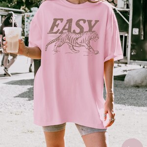 May include: A light pink t-shirt with the word "EASY" in brown block letters above a brown tiger illustration. The shirt is a loose fit and has short sleeves.