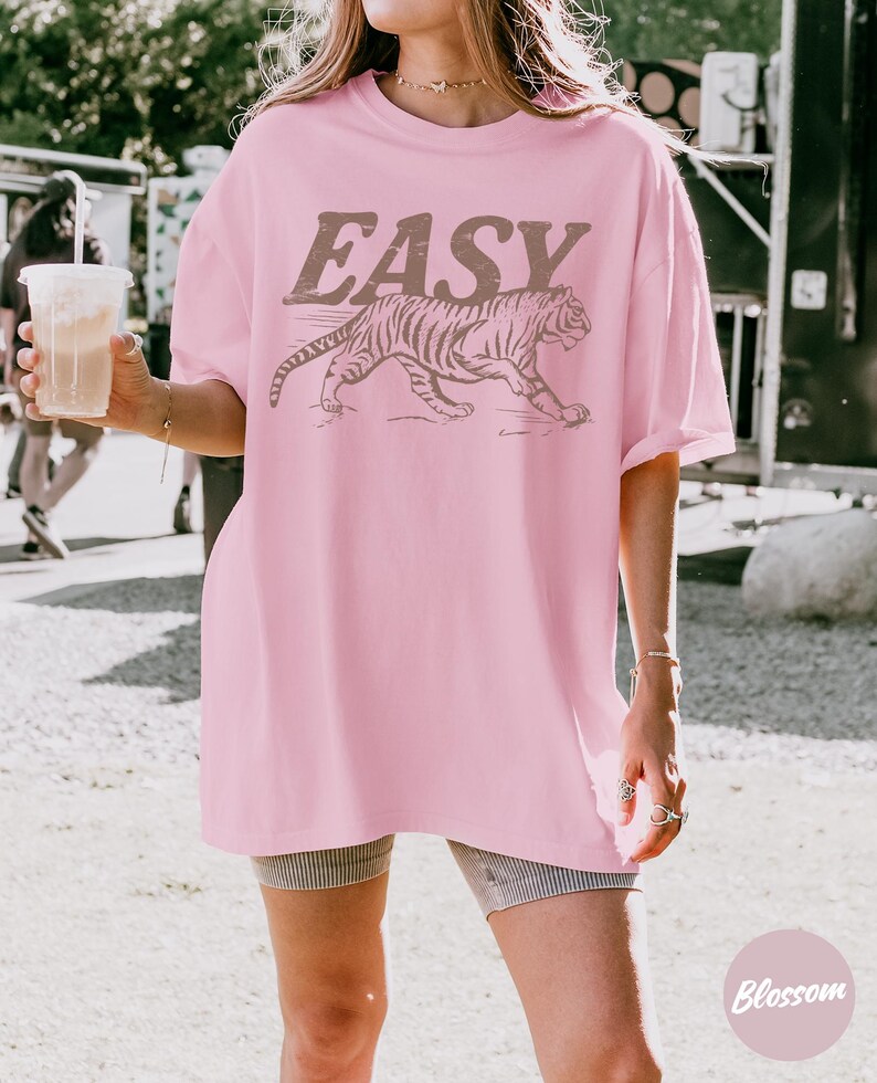 May include: A light pink t-shirt with the word "EASY" in brown block letters above a brown tiger illustration. The shirt is a loose fit and has short sleeves.