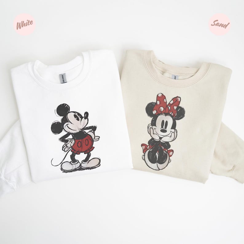 May include: Two sweatshirts, one white and one sand-coloured, each featuring a cartoon character. The white sweatshirt has a Mickey Mouse graphic in black, red, and white. The sand-coloured sweatshirt has a Minnie Mouse graphic in black, red, and white.
