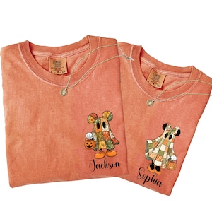 以下が含まれることがあります： オレンジ色のComfort ColorsのTシャツ2枚。漫画のゴーストのデザイン。1枚のシャツには「Jackson」という名前、もう1枚には「Sophia」という名前が書かれています。各ゴーストはジャック・オー・ランタンを持ち、パッチワークのキルトを着ています。金のネックレスがシャツにかけられています。