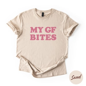 May include: Sand-colored t-shirt with the text "MY GF BITES" in pink, a playful design. The shirt is made of a soft material and has short sleeves. The color of the shirt is sand.