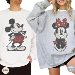 May include: Two sweatshirts featuring Disney's Mickey and Minnie Mouse. One is white with Mickey in red shorts, the other is grey with Minnie wearing a red bow. The sweatshirts are casual wear.