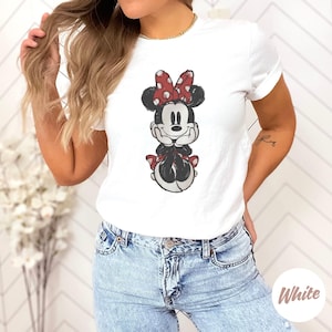 May include: White t-shirt with a graphic of Minnie Mouse. The cartoon character is in black, white, and red, with a red polka-dot bow. The shirt is worn with blue jeans.