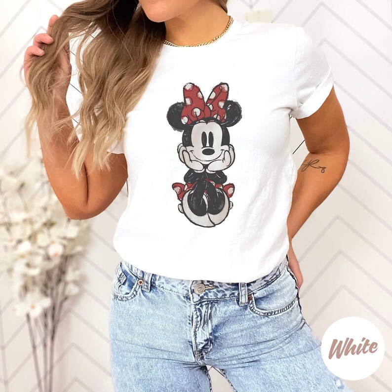 May include: White t-shirt with a graphic of Minnie Mouse. The cartoon character is in black, white, and red, with a red polka-dot bow. The shirt is worn with blue jeans.