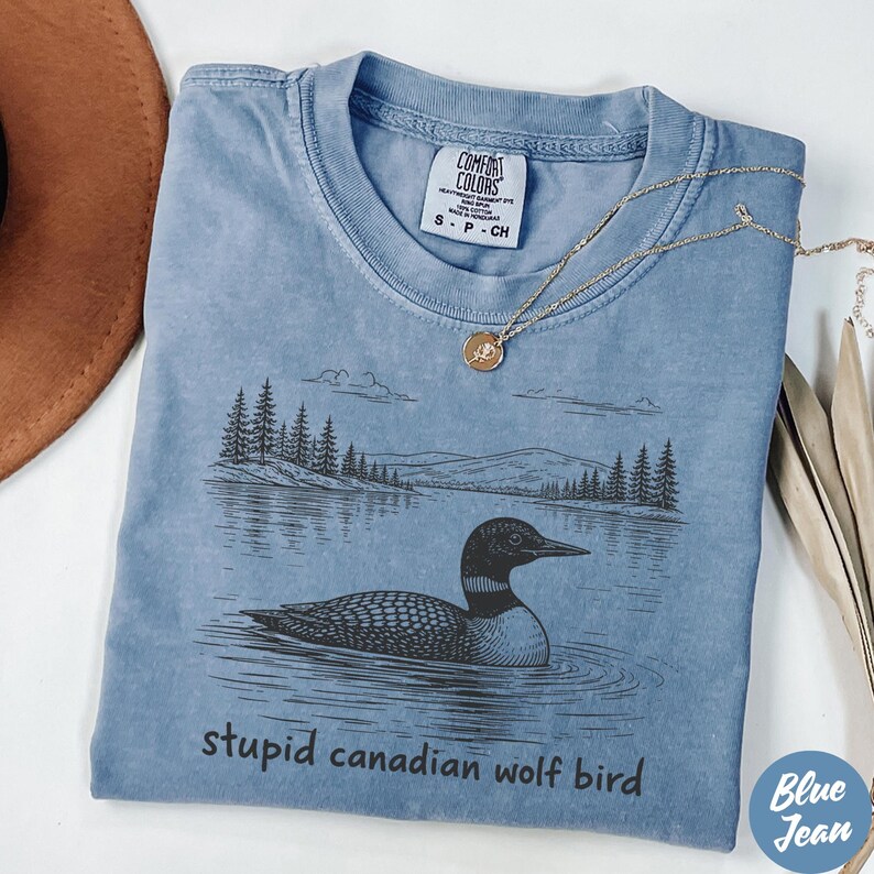 May include: A blue t-shirt with a graphic of a loon swimming on a lake, with trees and mountains in the background. The text "stupid canadian wolf bird" is below the graphic. A gold necklace is draped across the shirt.