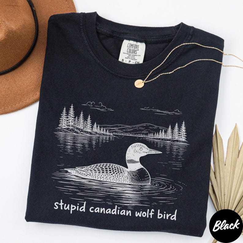 May include: Black t-shirt with a white graphic of a loon on a lake, with trees and mountains in the background. The text "stupid canadian wolf bird" is below the graphic. A gold necklace and a brown hat are also visible.