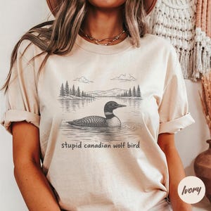 May include: Ivory-colored t-shirt featuring a detailed illustration of a loon swimming on a lake, with a forest and mountains in the background. The text "stupid canadian wolf bird" is printed below the image.