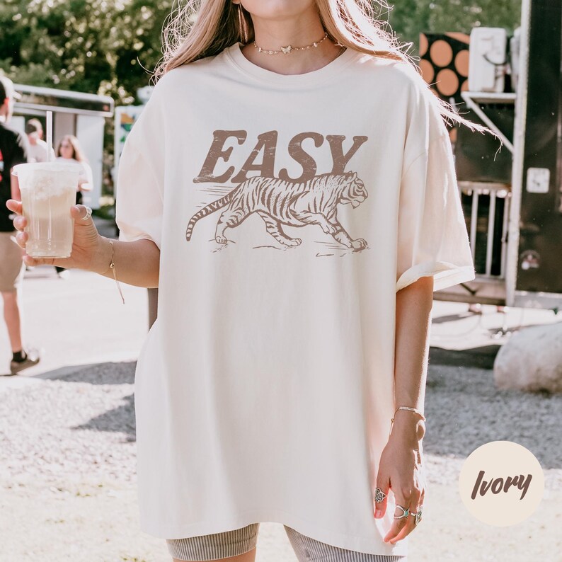 May include: Ivory-colored t-shirt with the word "EASY" in brown block letters above a brown tiger illustration. The shirt is a relaxed fit and has short sleeves. The word "Ivory" is written in cursive in the bottom right corner.