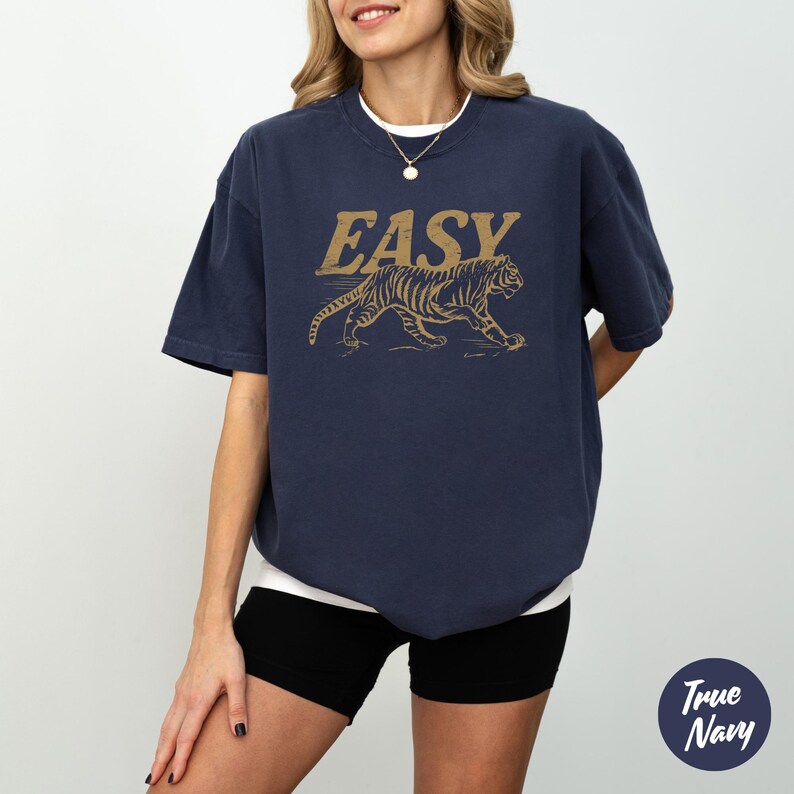 May include: Navy blue t-shirt with a gold tiger graphic and the word "EASY" above it. The shirt has short sleeves and a relaxed fit. The model is wearing black shorts and a white undershirt.