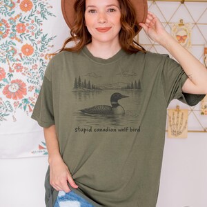 May include: Sage green t-shirt featuring a black and white illustration of a loon on a lake with trees and mountains in the background. The text "stupid canadian wolf bird" is below the graphic. The shirt is a relaxed fit.
