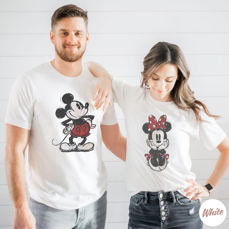 May include: Two white t-shirts featuring Mickey and Minnie Mouse. Mickey is depicted in red shorts and Minnie in a red polka dot bow. The shirts are worn by two people. The word "White" is in the bottom right corner.