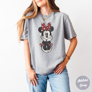 May include: Athletic grey t-shirt with a distressed Minnie Mouse graphic. Minnie is shown with a red bow with white polka dots, a red dress, and black shoes. The shirt is a casual, unisex style.