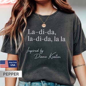 Puede incluir: Camiseta gris oscuro con el texto "La-di-da, la-di-da, la la" y "Inspired by Diane Keaton" en blanco. La camiseta es de la marca Pepper y se envía desde EE. UU.
