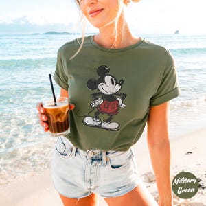 May include: Military green t-shirt with a distressed Mickey Mouse graphic in black, white, and red. The tee has short sleeves and a crew neck. The model is wearing denim shorts and holding a glass of iced coffee on a beach.