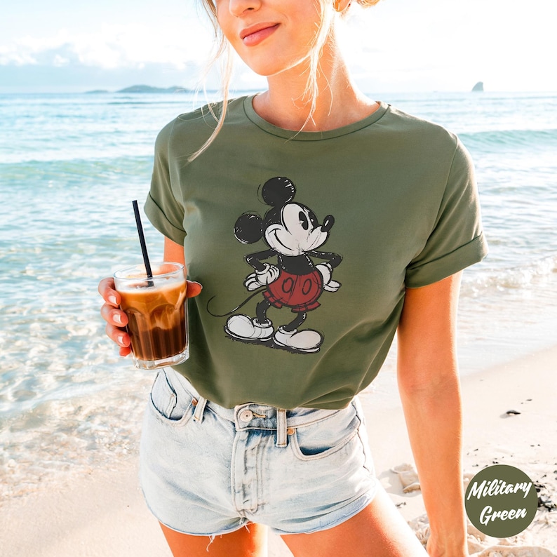 May include: Military green t-shirt with a distressed Mickey Mouse graphic in black, white, and red. The tee has short sleeves and a crew neck. The model is wearing denim shorts and holding a glass of iced coffee on a beach.