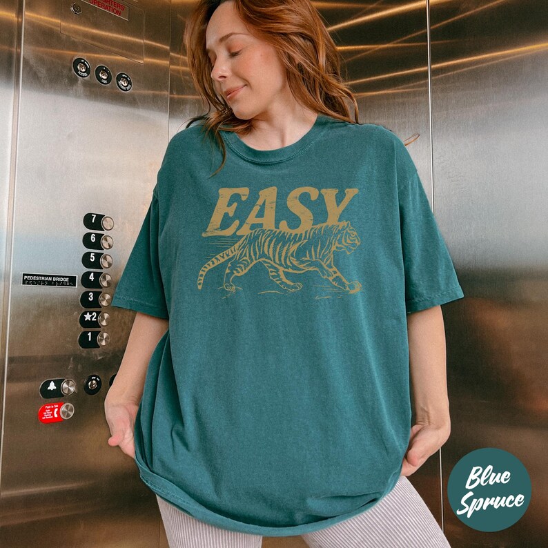 May include: Teal-colored t-shirt with the word "EASY" above a gold tiger illustration. The shirt has short sleeves and a relaxed fit. The text "Blue Spruce" is in a circle in the bottom right corner.