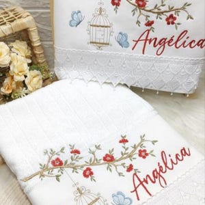 Puede incluir: Toallas blancas bordadas con dise&ntilde;os florales y de jaulas para p&aacute;jaros. Las toallas presentan flores rojas, mariposas azules y el nombre "Ang&eacute;lica" en escritura roja. El borde inferior tiene un ribete de encaje decorativo.
