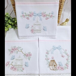Puede incluir: Mantitas de beb&eacute; blancas bordadas con dise&ntilde;os florales, jaulas y casitas para p&aacute;jaros. Cada manta presenta detalles en rosa y azul, delicados bordes de encaje y un lazo celeste. Las mantas est&aacute;n apiladas.