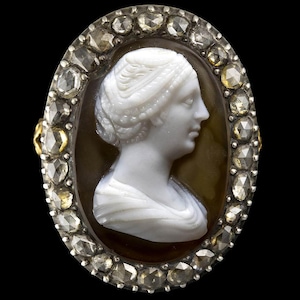 May include: Oval-shaped antique cameo ring featuring a white profile of a woman. The cameo is set in a brown background and surrounded by a silver frame with numerous clear and yellow-toned gemstones. The ring is set against a black background.