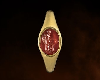 May include: A gold ring featuring a pointed, navette-shaped bezel set with a translucent red gemstone. The stone is intaglio-carved with the figure of a seated person holding a staff and a small circular object, set within a smooth, polished gold band.