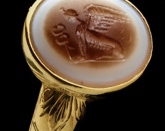 May include: A gold ring with a round, light-coloured stone featuring a brown, detailed carving of a winged figure. The ring's band has an ornate, leaf-like design. The ring is set against a black background.