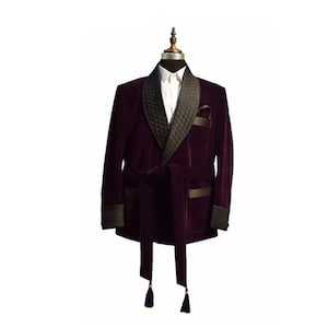May include: A burgundy velvet smoking jacket with a black quilted lapel and trim. The jacket features a matching belt and pocket square, and is displayed on a mannequin. The sleeves have black trim and the belt has tassels.