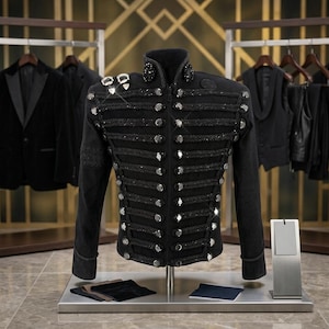 Può includere: Giacca nera in stile militare con bottoni argentati e finiture decorative in corda. La giacca presenta abbellimenti sulle spalle e sul colletto. Altri capi neri sono sullo sfondo.
