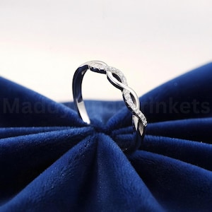 May include: A silver ring with an infinity symbol design. The ring has small, clear stones set along the infinity loop. The ring is displayed on a dark blue velvet surface.