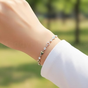 May include: A silver bracelet with heart-shaped charms and sparkling details. The bracelet is worn on a wrist, with a white sleeve visible. The background is a soft blur of green, suggesting an outdoor setting.